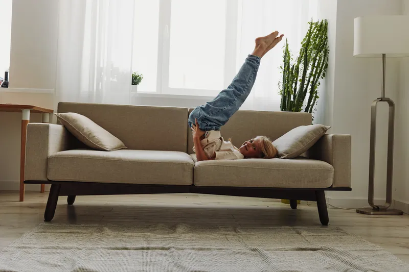 Young boy lying on modern sofa lifting legs up and playing with toy in bright living room with natural light.