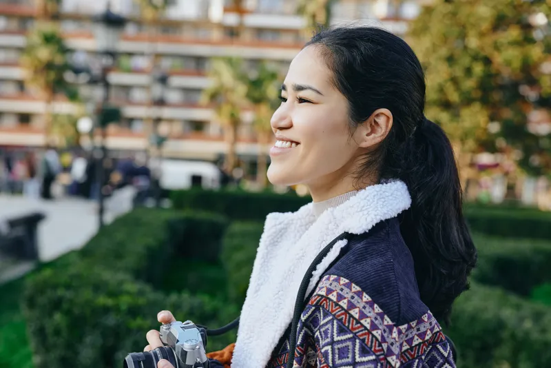 Smiling woman outdoors with a camera, wearing a jacket and scarf, in a park setting with greenery and urban buildings, candid lifestyle photography under warm sunlight, conveying leisure and cheerful