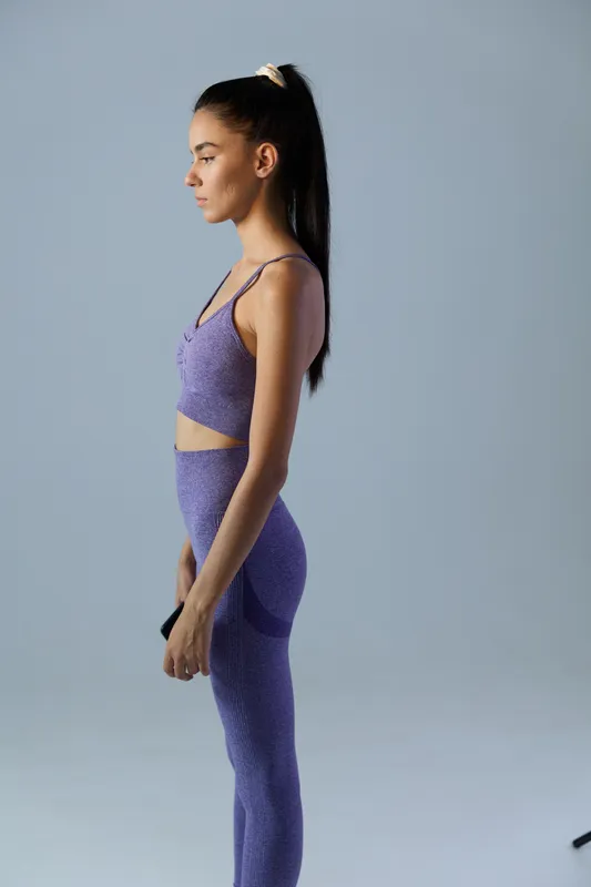 woman sport fitness in purple activewear standing sideways in studio with minimal gray background, focused expression, and long dark ponytail, healthy lifestyle concept
