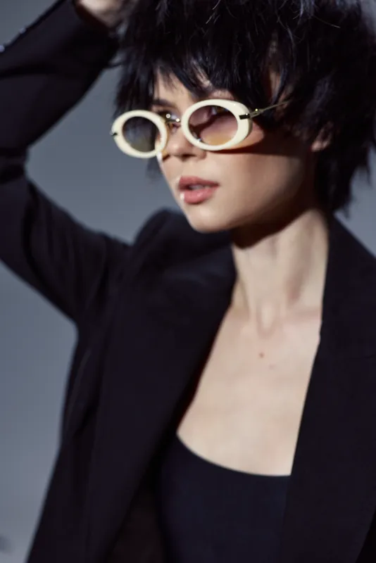 Stylish woman wearing white round sunglasses and a black blazer with short hair posing confidently indoors.