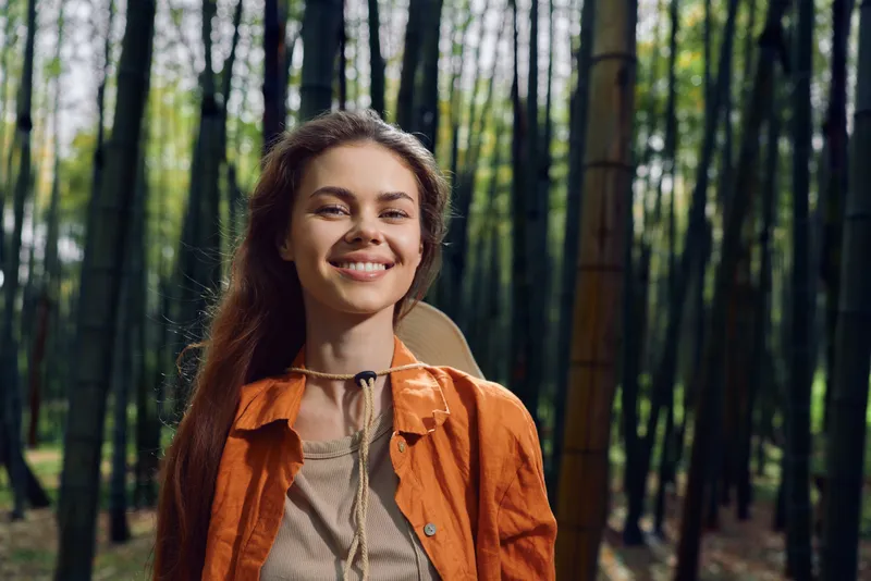 Woman smile forest bamboo backpack hiking nature portrait outdoor travel lifestyle young adult enjoying sunlight in green grove, casual orange jacket and relaxed happy expression.