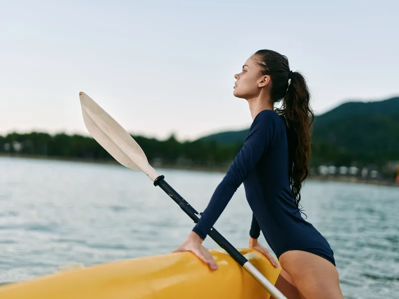 Woman with long hair wearing a navy swimsuit sitting on a kayak holding a paddle at calm lake during early morning light