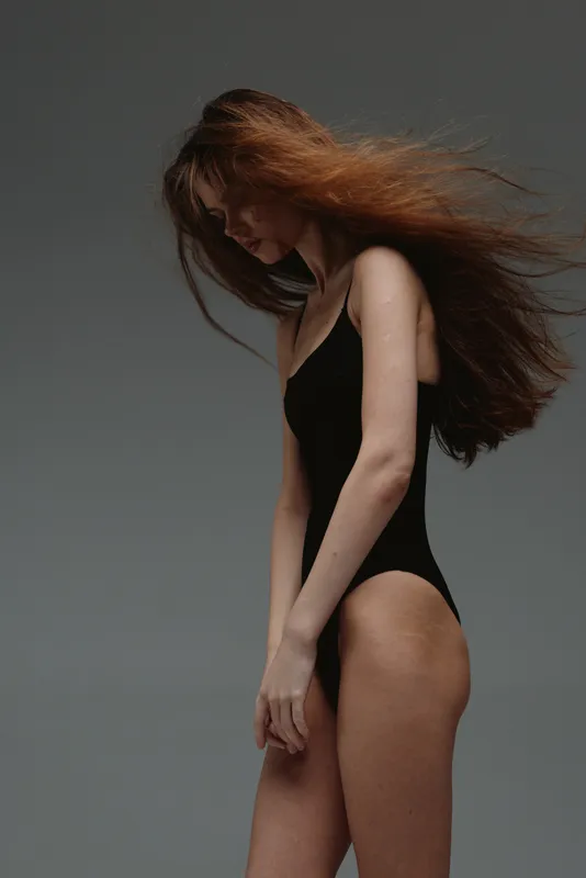 slender woman with long red hair wearing black swimsuit against gray background