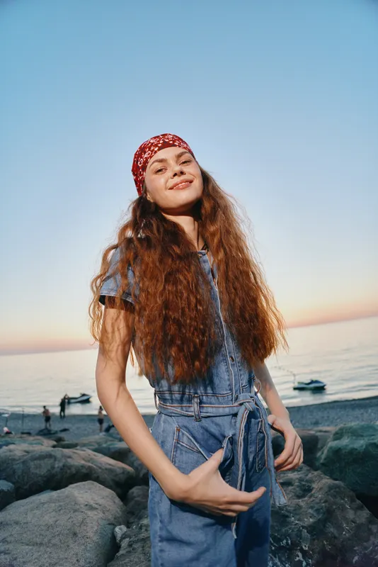 smiling woman standing on rocky beach wearing denim overalls and red headscarf at sunset with calm sea and boats in background