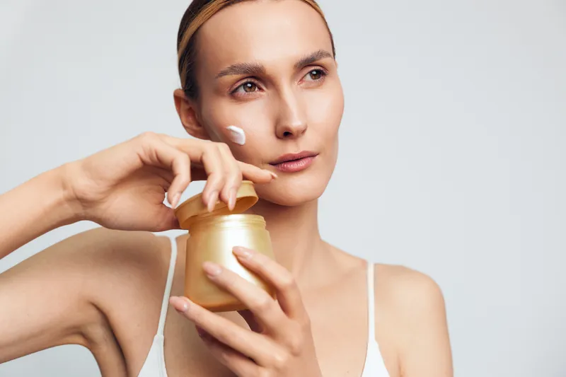 Young woman applying face cream on cheek, skincare routine, natural beauty, clean skin, focused expression, soft lighting, minimal makeup, white background, health care concept