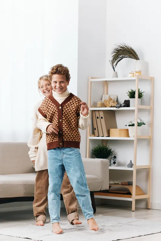 Authenticity shines in this lifestyle scene of children at play, wearing school inspired outfits and cozy knitwear as they pose with cheerful energy in a bright living room.
