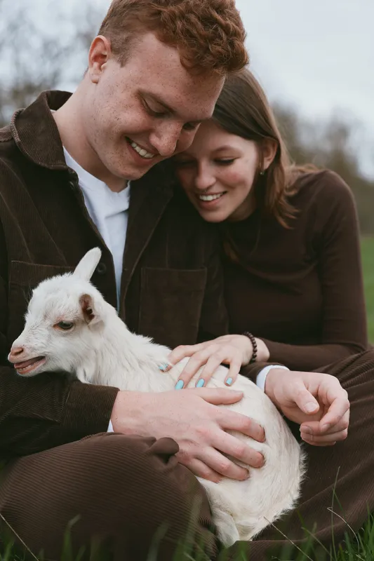Young couple sitting on grass gently holds a white baby goat together while sharing a joyful moment outdoors in casual autumn clothing.