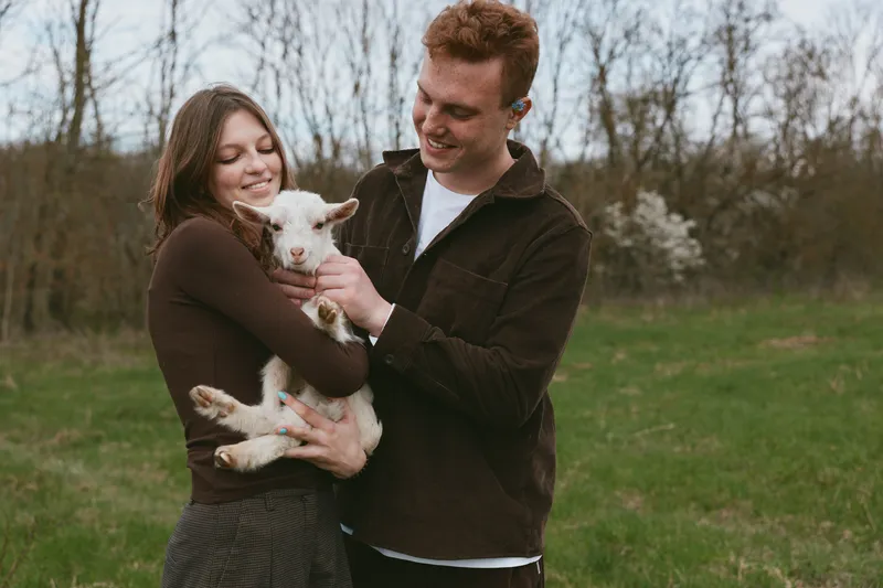 Young couple embraces a small white baby goat outdoors in early spring, sharing a joyful moment in casual dark clothing with natural background and soft daylight.