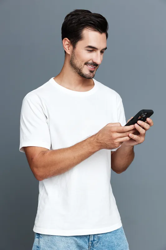 Smiling young man wearing white t-shirt stands against grey background looking at mobile phone with cheerful expression. Modern lifestyle concept.