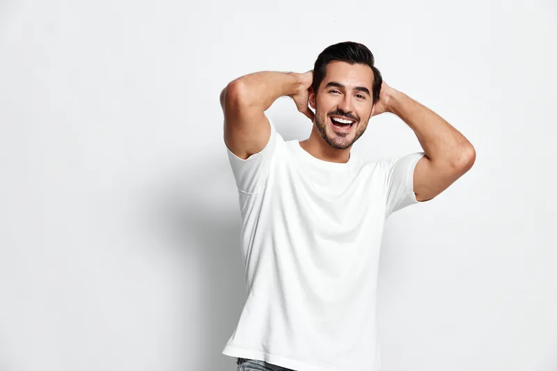 Smiling man with dark hair wears a casual white t-shirt and jeans, poses happily with hands behind head, isolated on plain light backdrop. Joyful lifestyle concept.