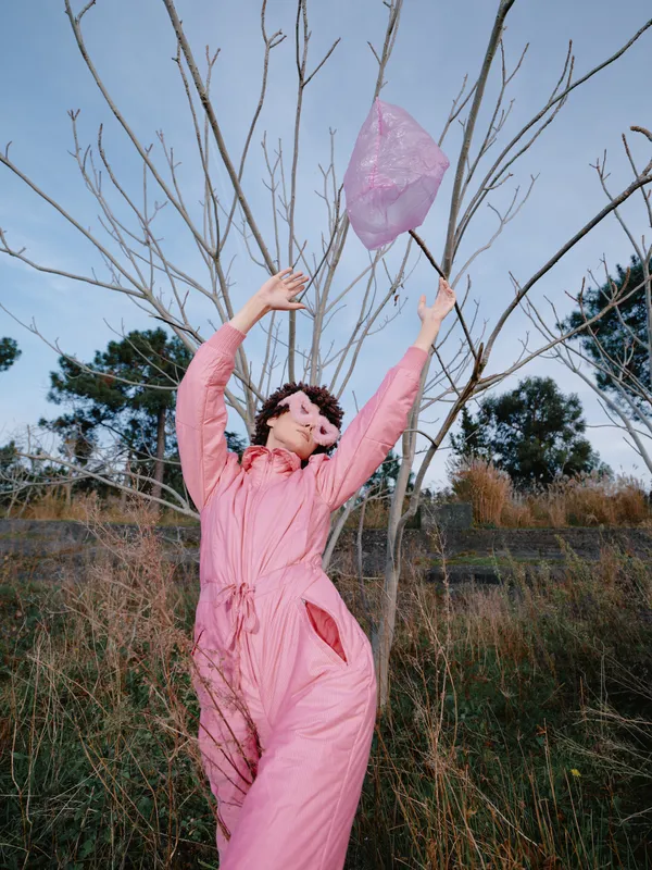 Outdoor scene with a woman in a pink jumpsuit reaching to release a pink balloon among bare trees, capturing a playful moment in a natural field setting on a breezy day and open space