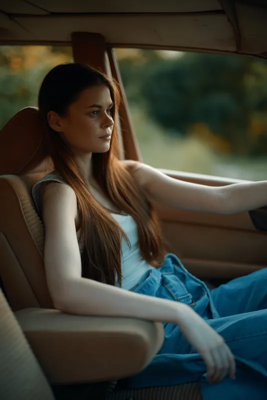 Young woman with long hair sitting in a stylish car, wearing casual clothes, looking pensive with a serene expression in a warm, natural setting.
