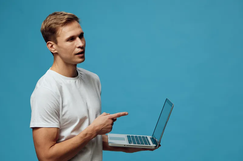 Young man with fair hair wearing casual white t-shirt holding open laptop and pointing at screen isolated on vibrant blue background in studio portrait. Technology and communication concept