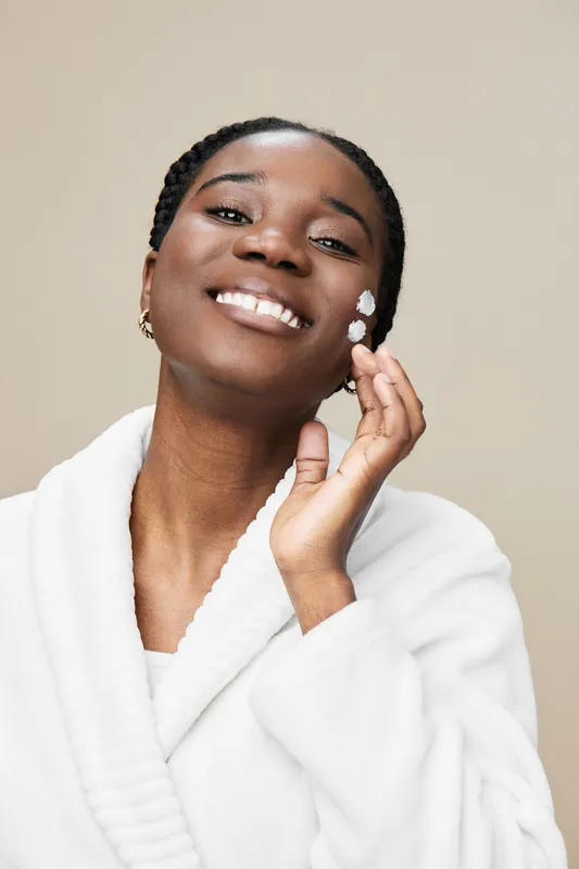 Smiling Black woman applying skincare in a cozy white robe against a soft neutral background, radiating confidence and showcasing a healthy skincare routine