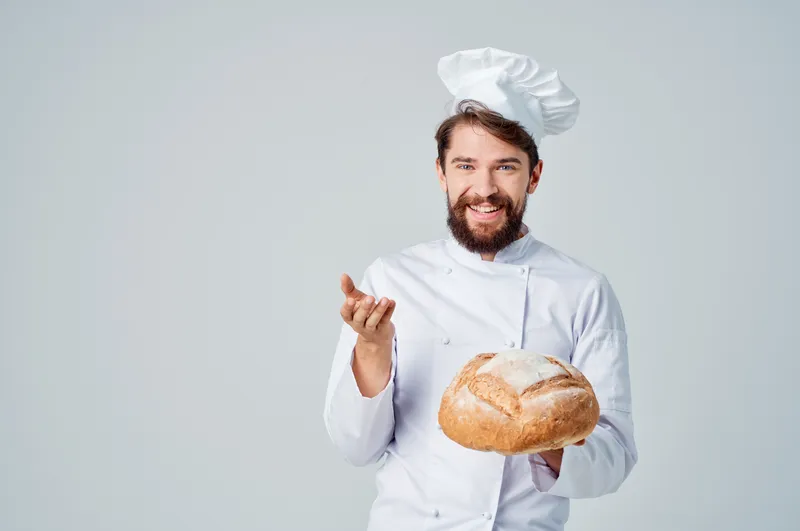 Бородатый мужчина-повар работает на кухне, рядом с ним хлебобулочные изделия на изолированном фоне