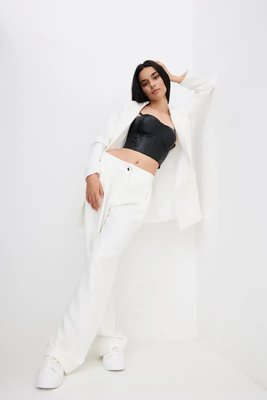 Stylish woman in white pantsuit and black corset top poses confidently against minimalist white background, showcasing modern fashion and youthful elegance.