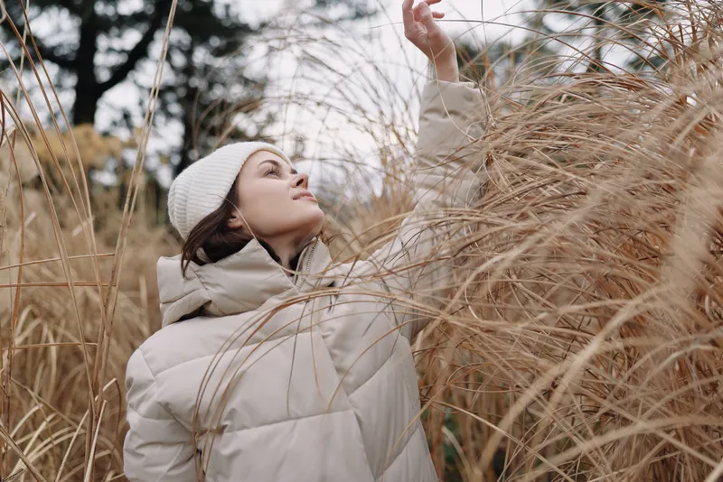 Woman in a warm coat and beanie reaches through tall dried grasses in a serene outdoor winter scene, capturing quiet mood in light breeze.