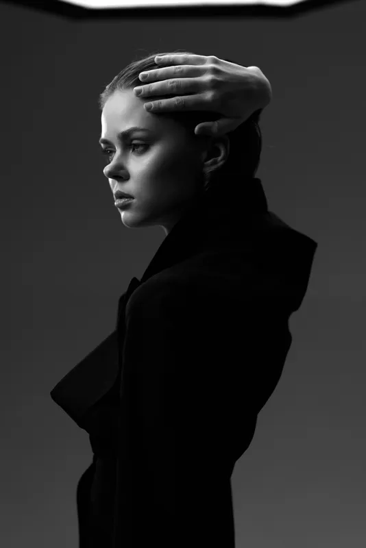 Black and white portrait of a thoughtful woman in dark clothing with hand on head, expressing contemplation and elegance.