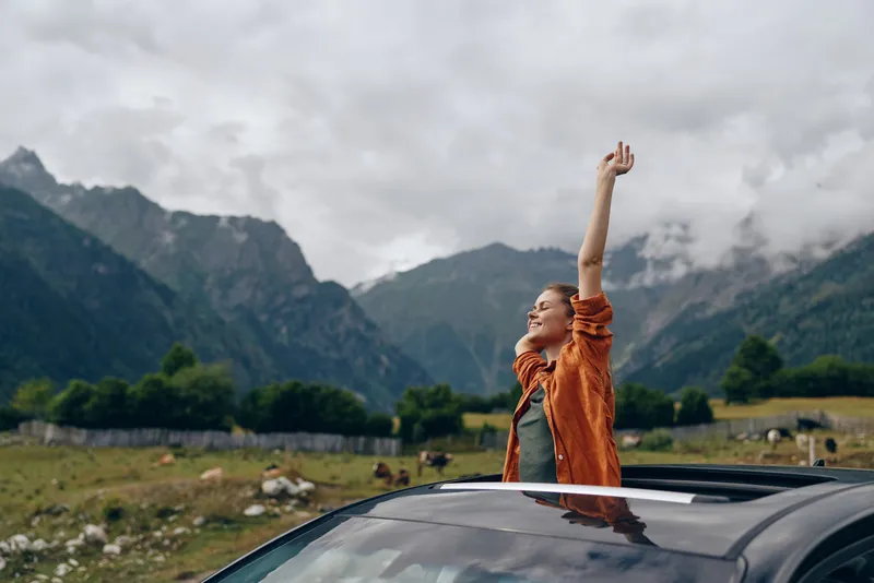 Joyful woman stands through a car sunroof raising her arm in celebration against a rugged mountain landscape outdoors, capturing a moment of freedom and adventure.