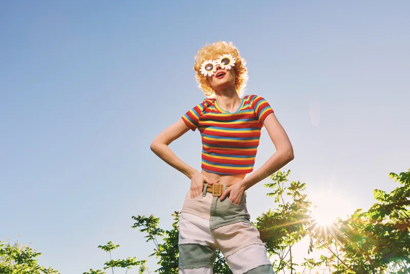 Smiling woman in a striped top and flower sunglasses poses outdoors under a bright sky, vibrant retro film colors creating a warm, cheerful lifestyle mood and carefree energy.