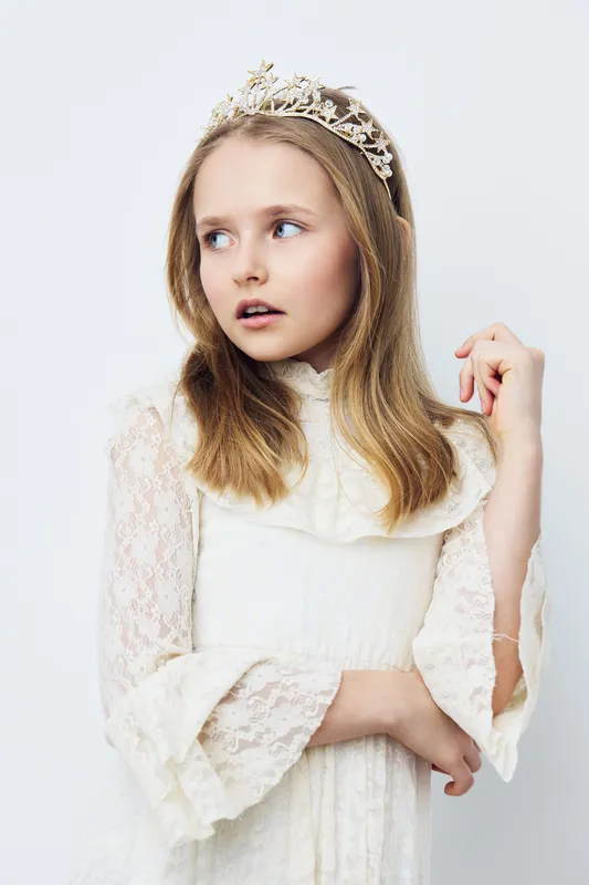 A young girl wearing a white lace dress and a crown with an elegant expression, symbolizing innocence and royalty in a soft, minimalist setting