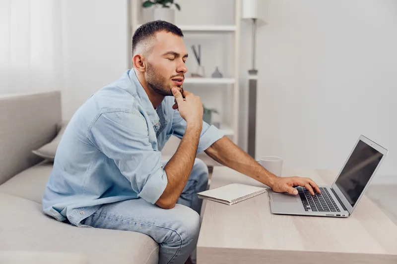 Thoughtful man working on a laptop at home, dressed casually in a light blue shirt. The cozy atmosphere enhances creativity and focus during the brainstorming session.