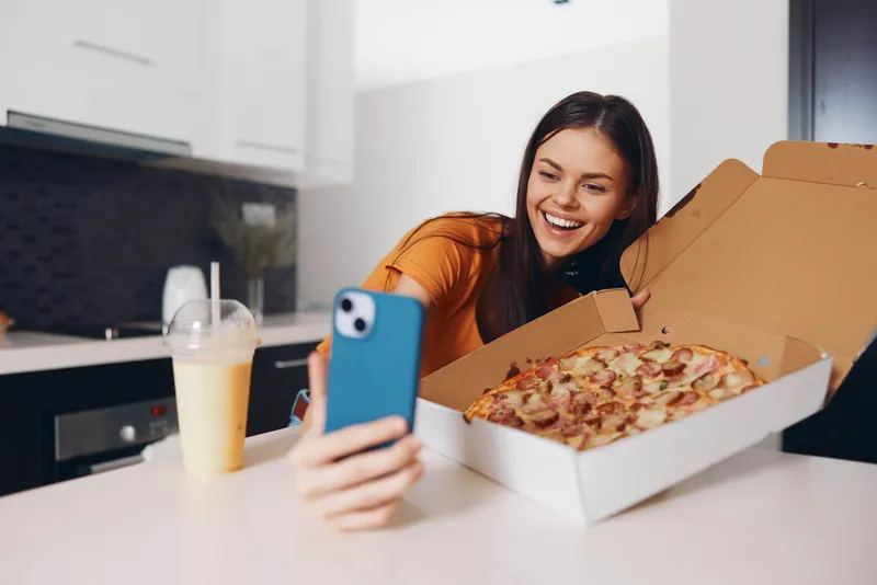 Женщина фотографирует на камеру телефона аппетитную домашнюю пиццу, стоящую на кухонном столе
