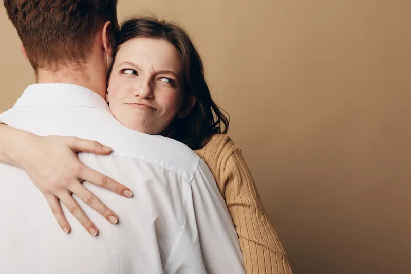 Young woman in brown sweater hugs man in white shirt with skeptical expression on beige background. Concept of mixed emotions, relationship tension, and complicated feelings.