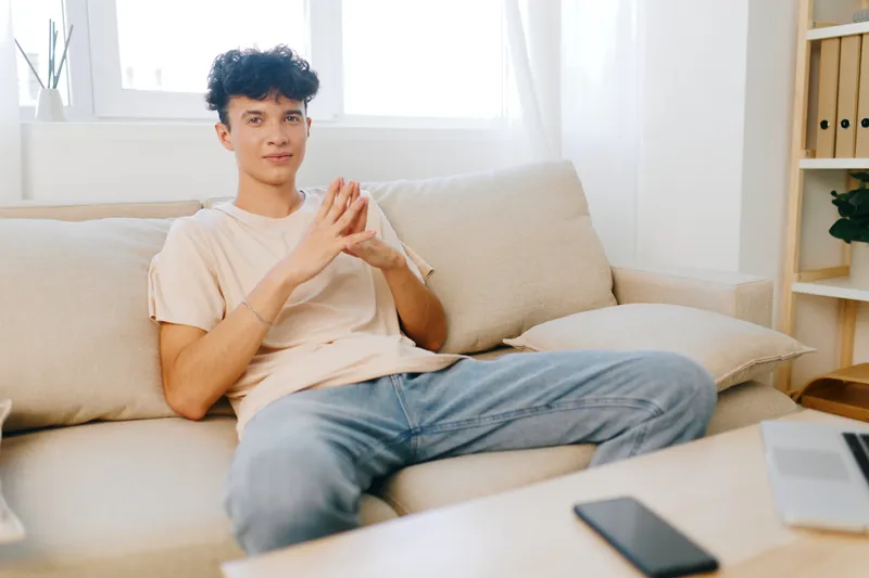 Молодой человек задумчиво сидит на удобном диване в светлой и уютной домашней обстановке