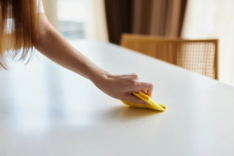 Cleaning hand cloth table wipe surface home household routine, female hand wiping white dining table with yellow cloth, closeup of domestic cleaning and tidy living space.