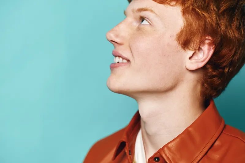 Profile of young redhead man smiling and looking up on blue background. Casual clothing with clear skin and joyful expression in studio lighting portrait photo.