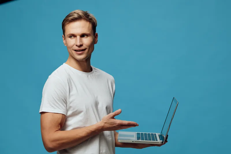 Smiling young man with short brown hair wearing casual white t-shirt holding open laptop in hand, looking at camera, isolated on plain turquoise background, studio portrait, people lifestyle concept, 