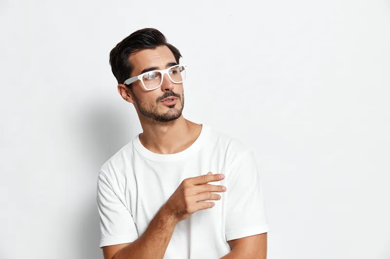 Stylish man in casual white t-shirt and glasses looking thoughtfully to the side, creating a contemplative mood against a light grey background.