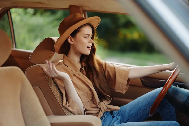 Stylish woman in a brown hat expressing emotions while driving a vintage car in nature, capturing a carefree summer vibe with warm colors.