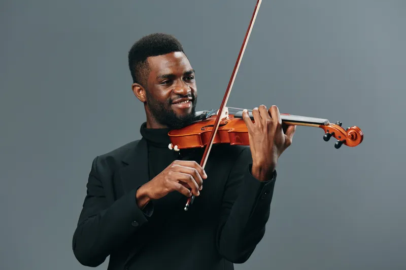 Элегантный афроамериканский мужчина в черном костюме играет на скрипке на сером фоне для концепций музыки и выступления