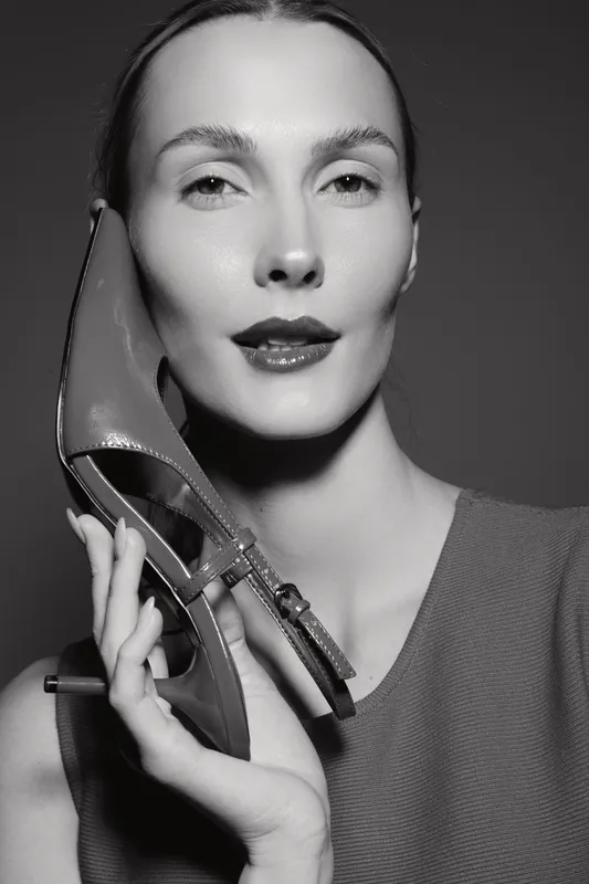 White woman with fair skin holds high heel close to face. Studio portrait in monochrome. Concept of fashion, elegance, and style.