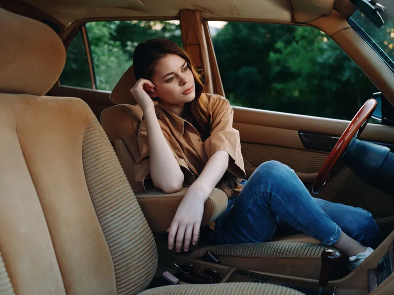 Young woman sitting in a vintage car, wearing a brown shirt and jeans, looking contemplative against a green background in a relaxed, nostalgic atmosphere.