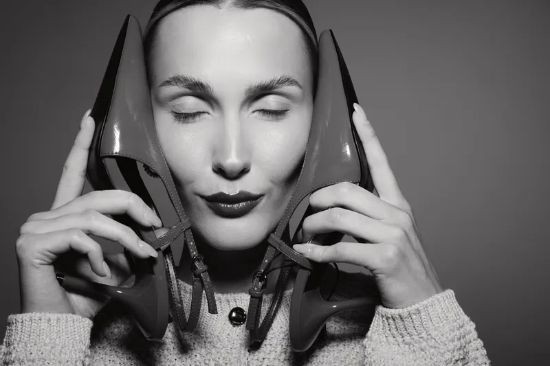 Young caucasian woman with fair skin holds high heel shoes near face with closed eyes in black and white photo. Concept of fashion and style expression.
