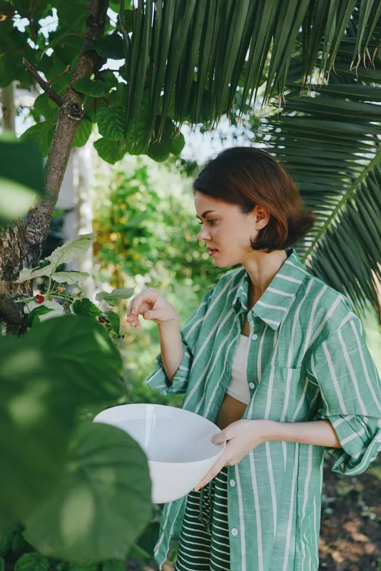 A young woman in a green striped shirt holds a white bowl while tending tropical plants in a sunlit garden.