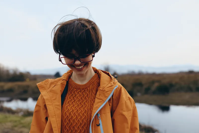 Woman lifestyle portrait outdoors beside river in autumn, orange sweater and coat, glasses, brunette hair. She smiles looking down, relaxed pose, countryside landscape with daylight and calm sky