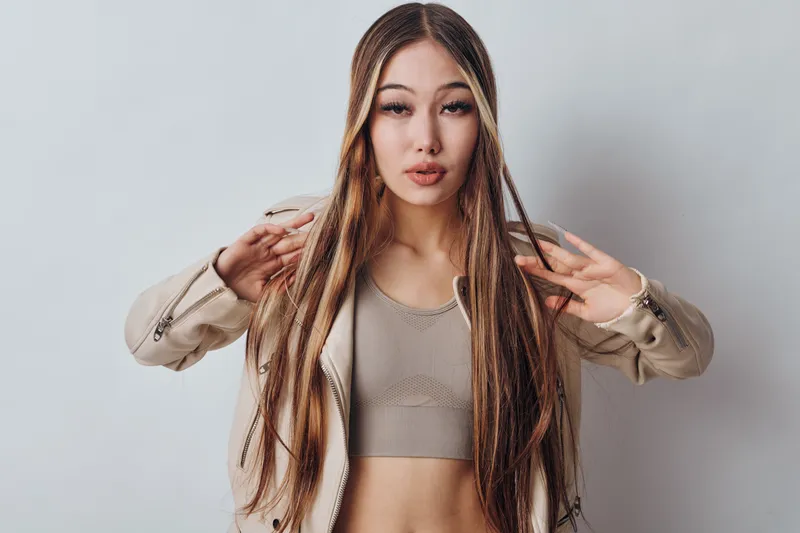 Young woman with long braided hair posing in beige jacket and crop top against white background, looking confidently at camera with stylish expression.