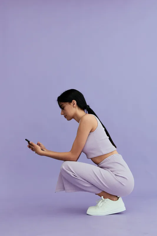 Young woman crouching and looking at smartphone, dressed in casual striped outfit and white sneakers, on soft purple background with relaxed mood.