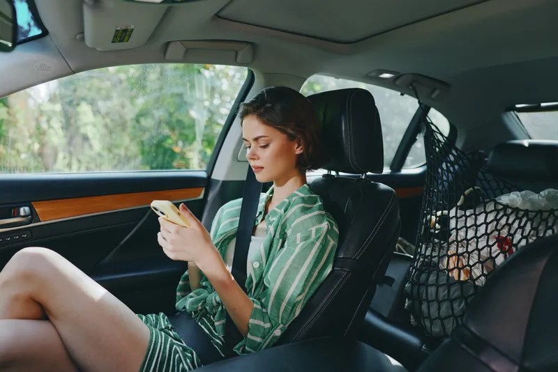 A young woman sits in a car backseat, absorbed in her phone, wearing a striped green shirt and relaxed pose.