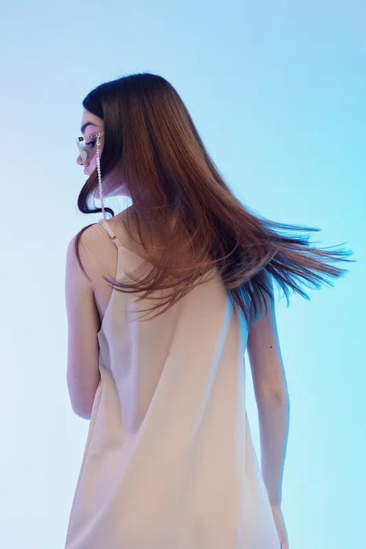 Young woman with long brown hair wearing a beige dress and sunglasses stands against a blue background. Concept of beauty, fashion, and style in studio lighting.