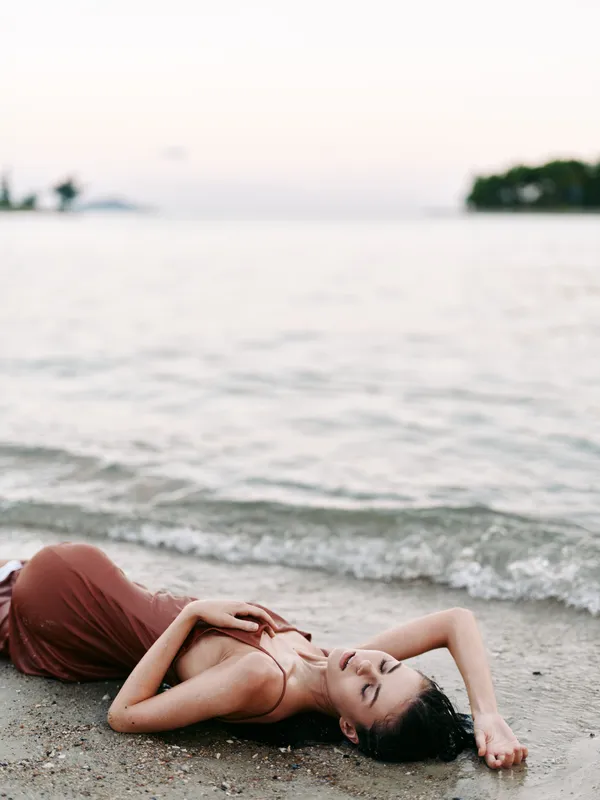 A tranquil beach scene features a relaxed woman in a brown dress lying on the sandy shore, delicate pose, gentle waves, soft light, serene setting.