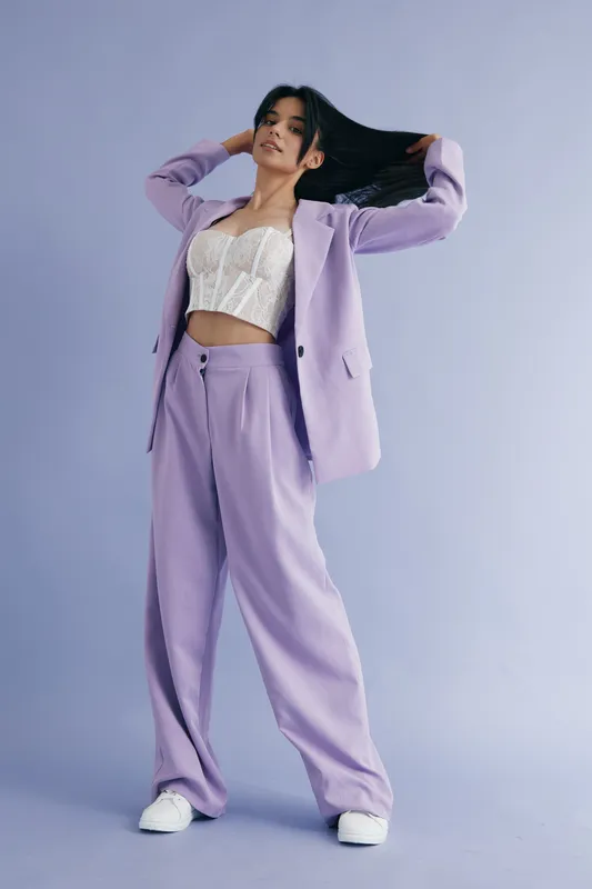 Stylish woman in lilac suit and white lace top posing confidently against soft lavender background, showcasing modern fashion and relaxed elegance in studio setting.