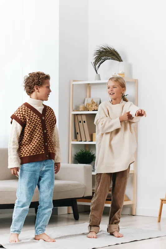 Two children wearing cozy sweaters stand in a bright living room, smiling and dancing as warm light floods the space and a soft rug anchors their playful movement.