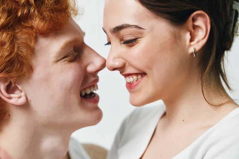 Couple smiling closely with joyful expressions in casual clothing on bright background. Young man and woman showing affection and happiness in intimate moment with natural light.