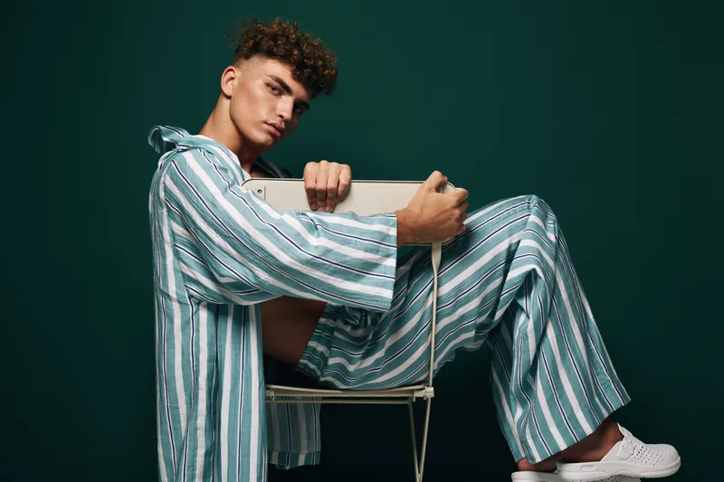 Young man in striped pajamas sitting on chair against dark background, casual pose with serious expression, fashion and lifestyle concept with relaxed mood.