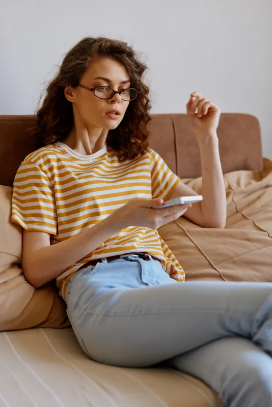 Woman with glasses sits on a sofa at home, wearing a striped shirt and jeans, focused on a tablet. A casual lifestyle moment in a cozy living space.